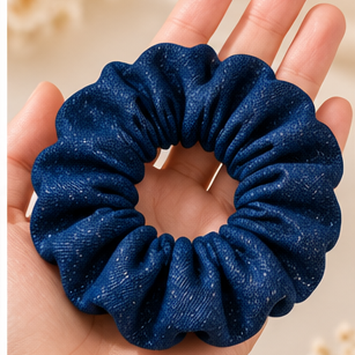 A dark blue fabric scrunchie with subtle white speckles, held in a person's hand against a soft beige background.