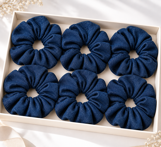 Six dark blue fabric hair scrunchies arranged in a 2x3 grid inside a cream-colored gift box, with soft decorative flowers in the corners.